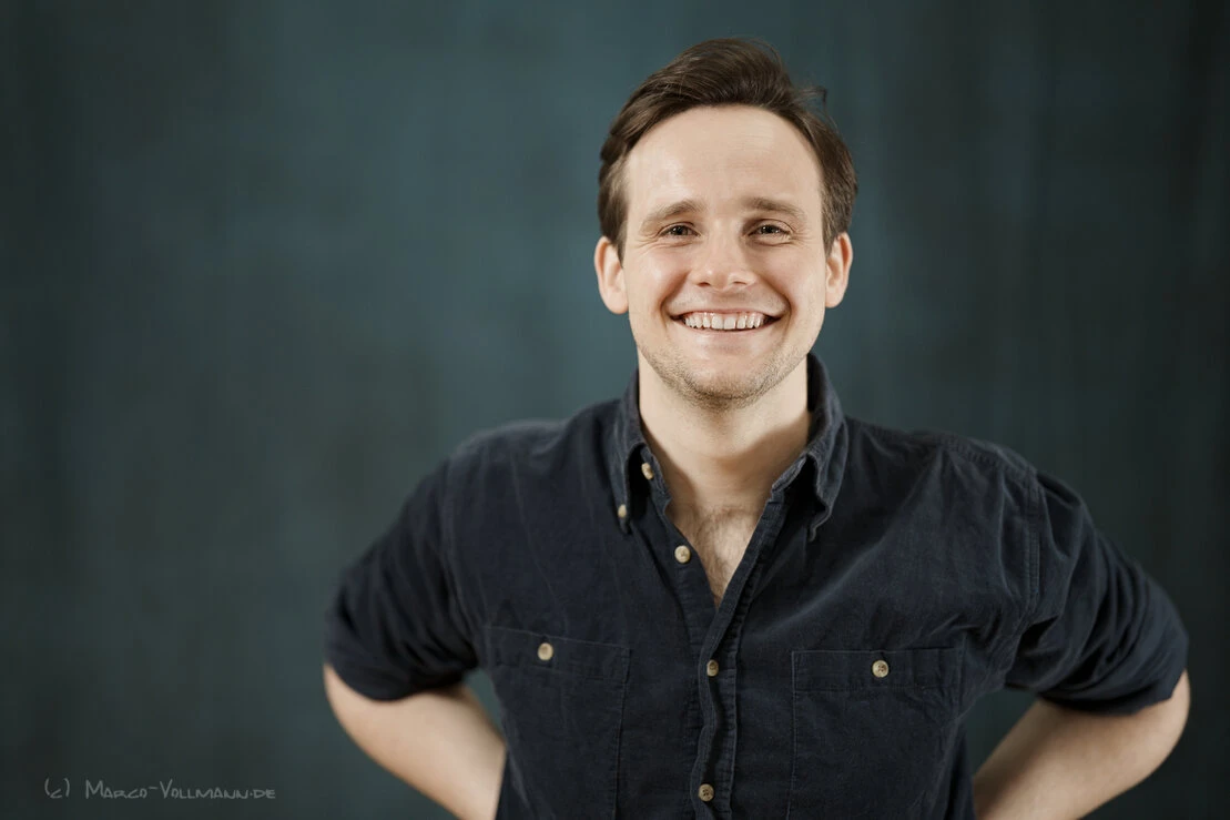 Junger Schauspieler in blauem Hemd mit in die Hüften gestützten Händen schaut lächelnd in Richtung des Betrachters