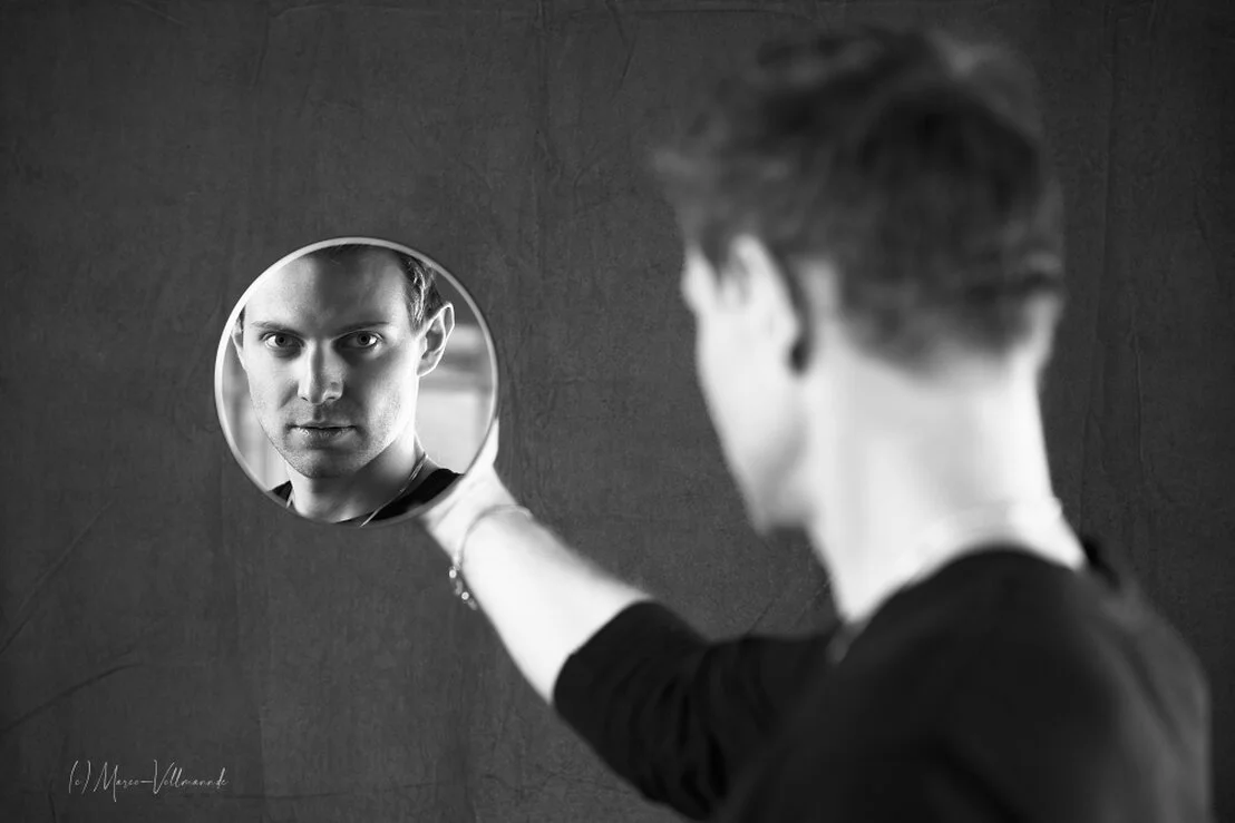 Schwarz-Weiß-Bild eines jungen Mannes, der durch einen Spiegel in der Hand in die Kamera sieht.